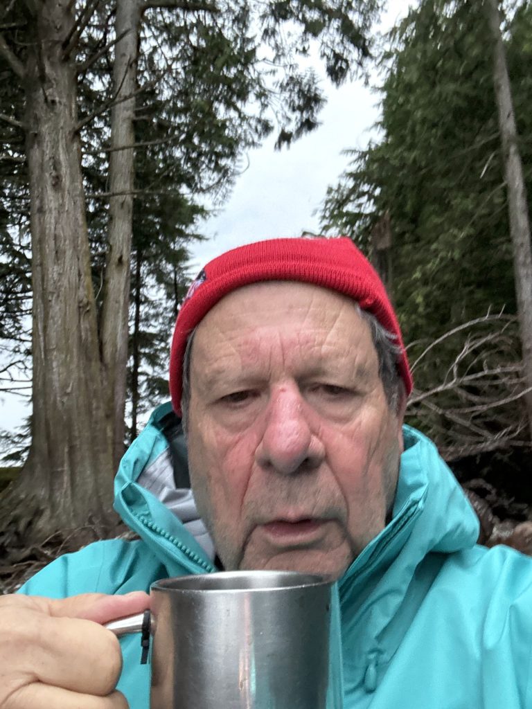 Bob Rein Reflections Off The Grid man with a rugged complexion wearing a red beanie and turquoise waterproof jacket, holding a stainless steel cup, with a dense forest background