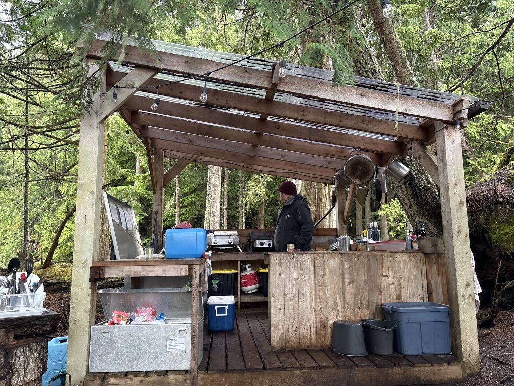 Bob Rein Reflections Off The Grid Rustic outdoor kitchen in a forest with wooden gazebo, metal roof, and a person in casual outdoor clothing standing inside