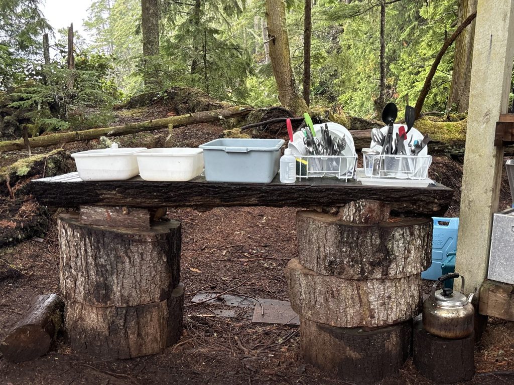 Bob Rein Reflections Off The Grid Rustic outdoor setup in a forest with wooden table made from tree trunks, featuring plastic containers and metal dish rack with utensils