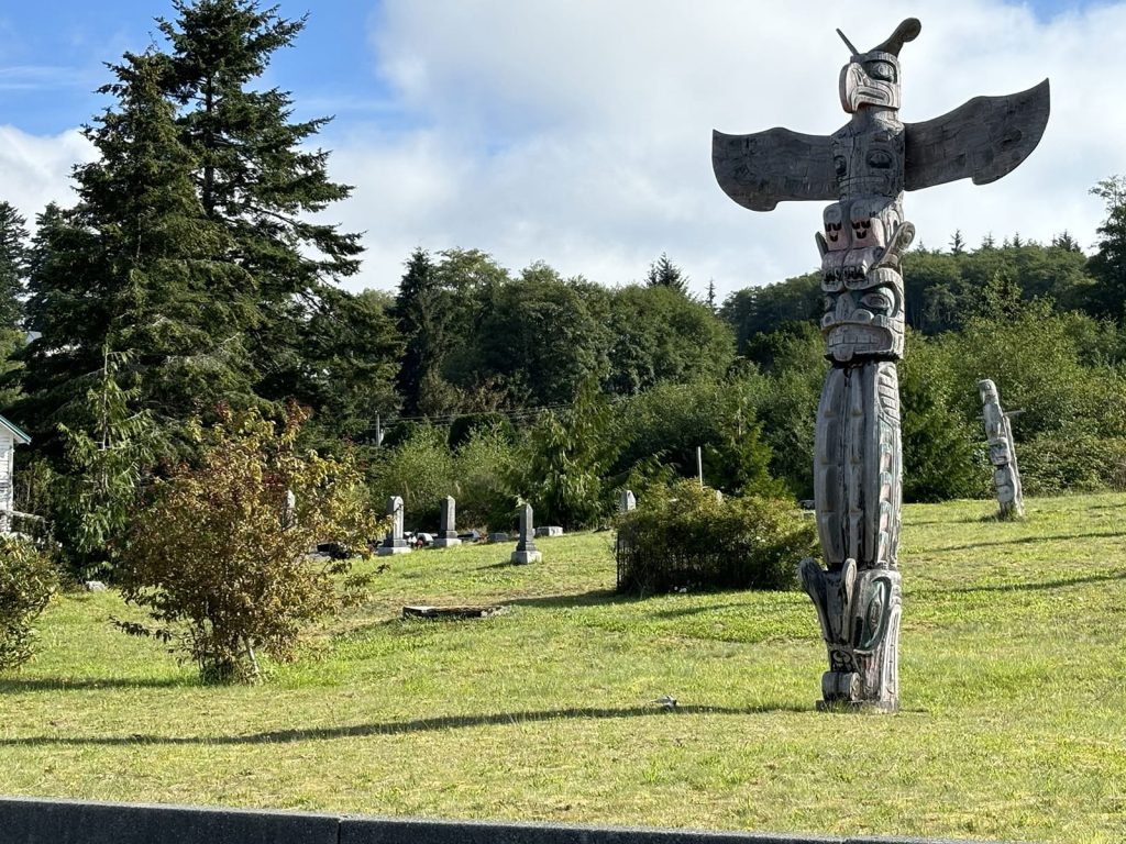 Bob Rein Reflections Off The Grid traditional totem poles in grassy area with carvings cemetary surrounded by forest and clear sky