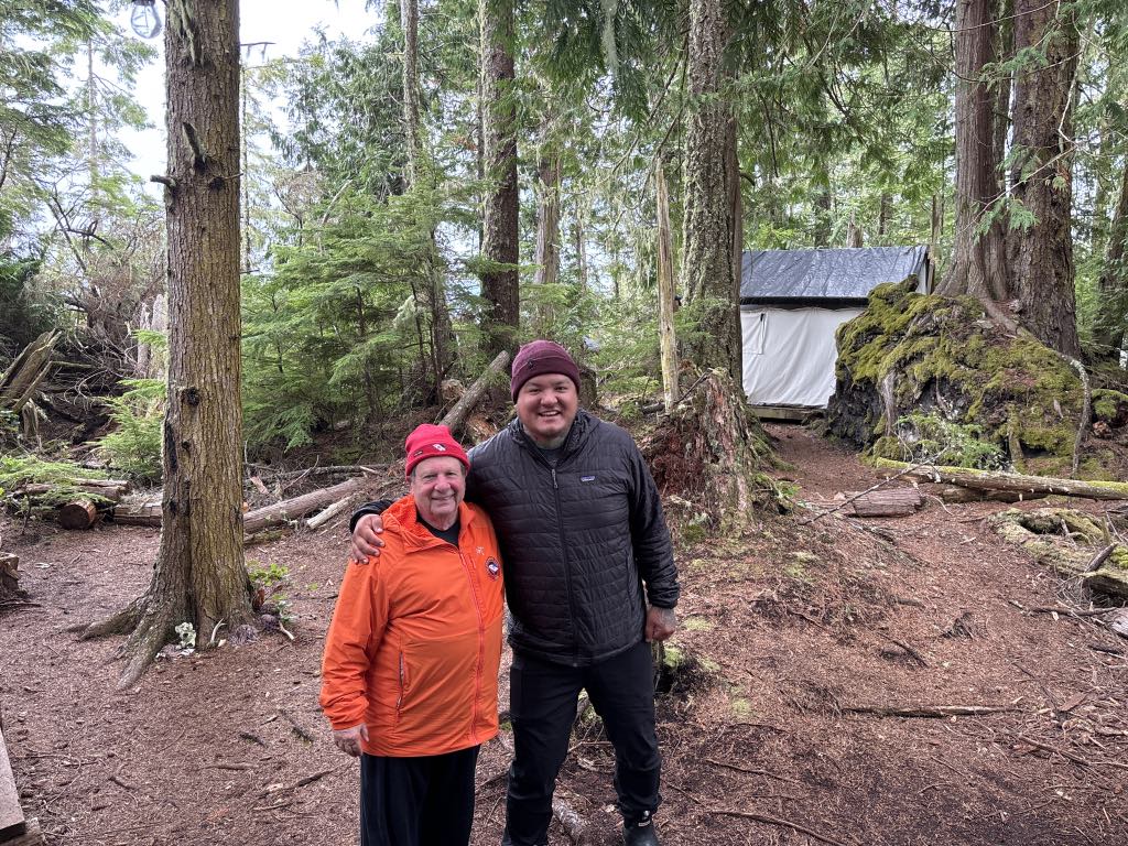 Bob Rein standing in the woods together smiling for a picture with a cabin in the background